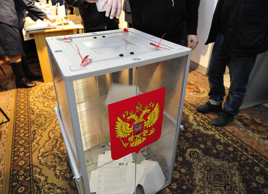 Transparent ballot box bearing the coat of arms of the Russian Federation, with four red security seals on the lid.