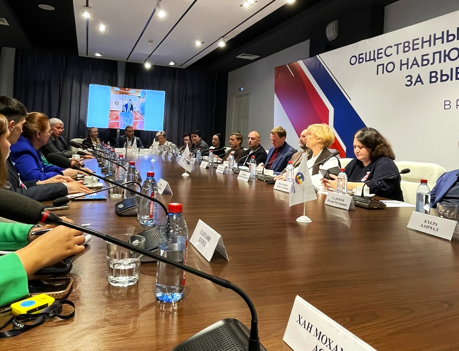 Meeting table with authorities, observers, and candidates, and a sign of the public observation headquarters in the background.