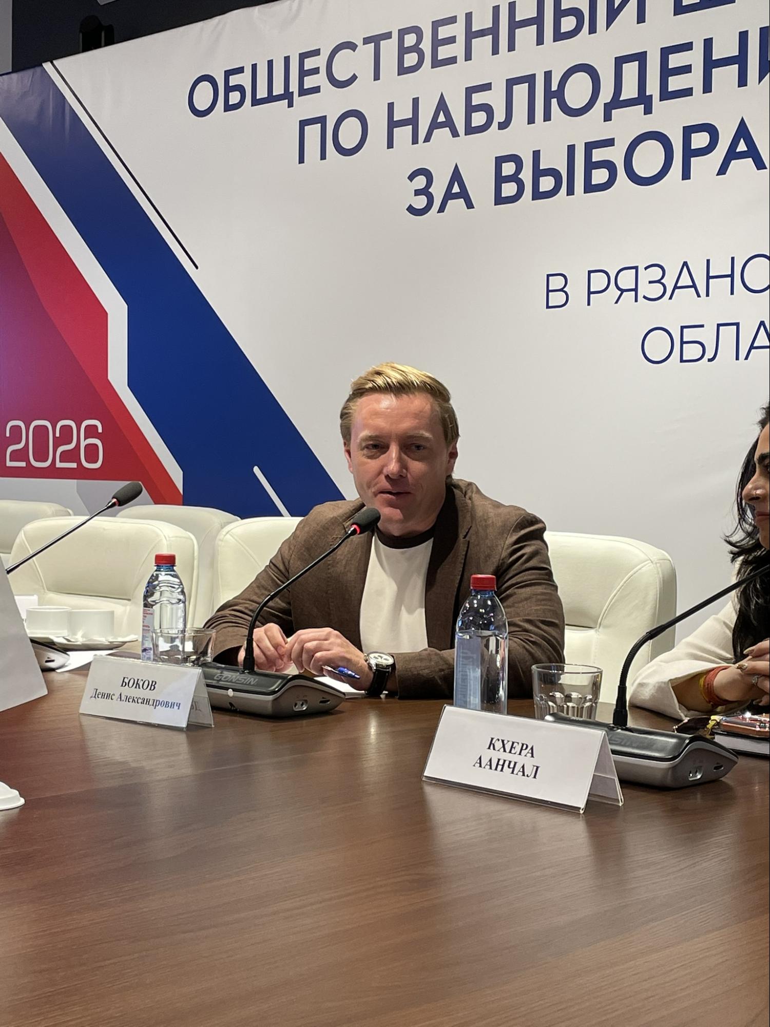 Denis Bokov speaking into a microphone, with the Public Election Observation Headquarters sign in the background.